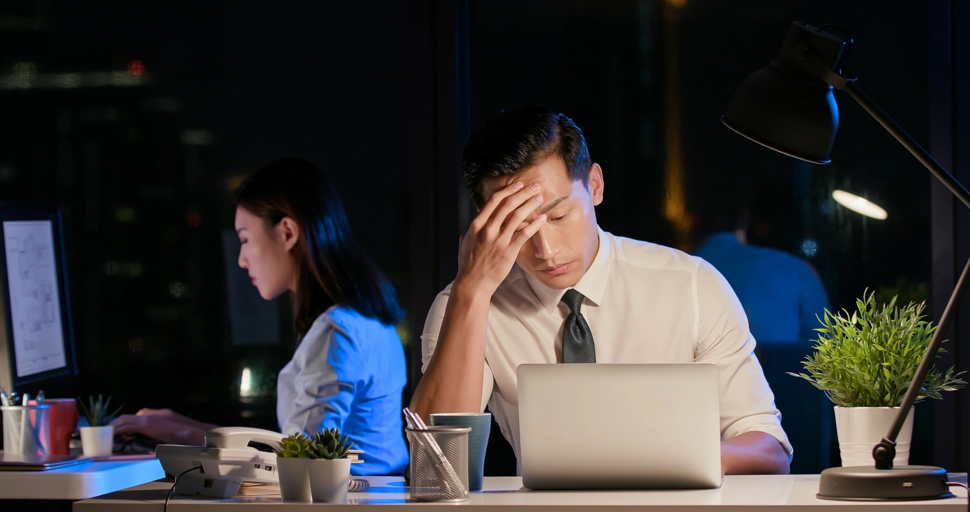 「燃え尽き症候群」のイメージ_暗い仕事場で頭を抑えながら仕事をする男性